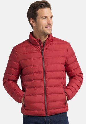 Mężczyzna w czerwonej pikowanej kurtce na zamek, z rękami w kieszeniach, patrzący w bok na jednolitym tle.