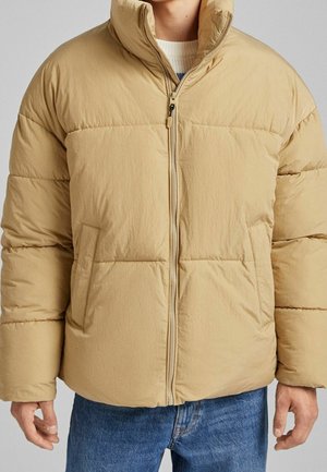 Veste matelassée beige avec un col haut, une fermeture éclair devant et deux poches latérales. Elle présente un design matelassé et une finition lisse et texturée.