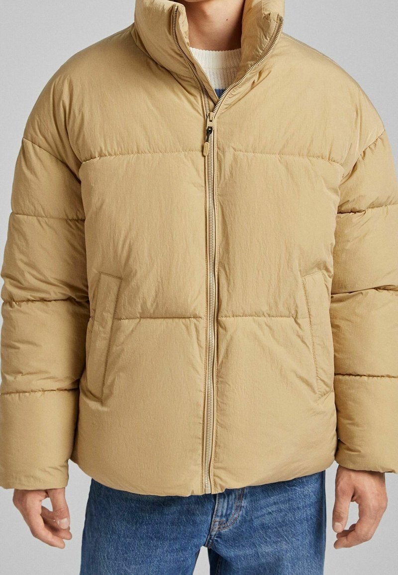Béžová péřovka s vysokým límcem, předním zipem a dvěma bočními kapsami. Má prošívaný design a hladký, strukturovaný povrch.