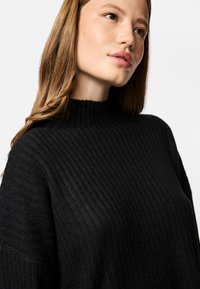 Jauna sieviete ar gaiši brūniem matiem valkā ribotu melnu džemperi ar augstu apkakli, skatoties nedaudz uz augšu un sānis pret baltu fona.