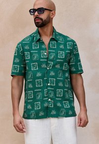 Green short-sleeve shirt with white abstract frame and leaf patterns. Lightweight fabric, button-front design, paired with light trousers.