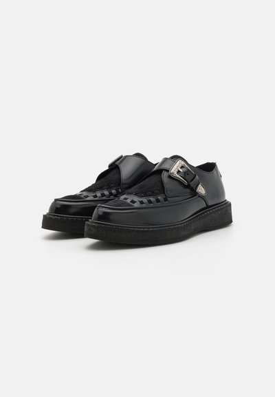 Chaussures en cuir noir avec un détail tressé et une boucle sur le côté. Elles disposent d'une semelle en caoutchouc plate avec un bord texturé et une doublure intérieure douce.