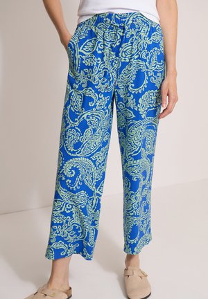 Persona con pantalones azules de estampado paisley, manos en los bolsillos, top blanco y zapatos beige sin cordones, de pie sobre un fondo liso.
