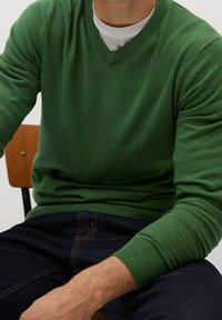Homme portant un pull vert à col en V par-dessus un t-shirt blanc, assis sur une chaise en bois avec un jean bleu foncé.