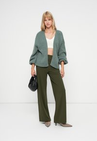 Cardigan spesso di colore verde chiaro, top bianco corto, pantaloni a gamba larga color oliva e borsa nera. Tacchi a punta beige completano l'outfit.