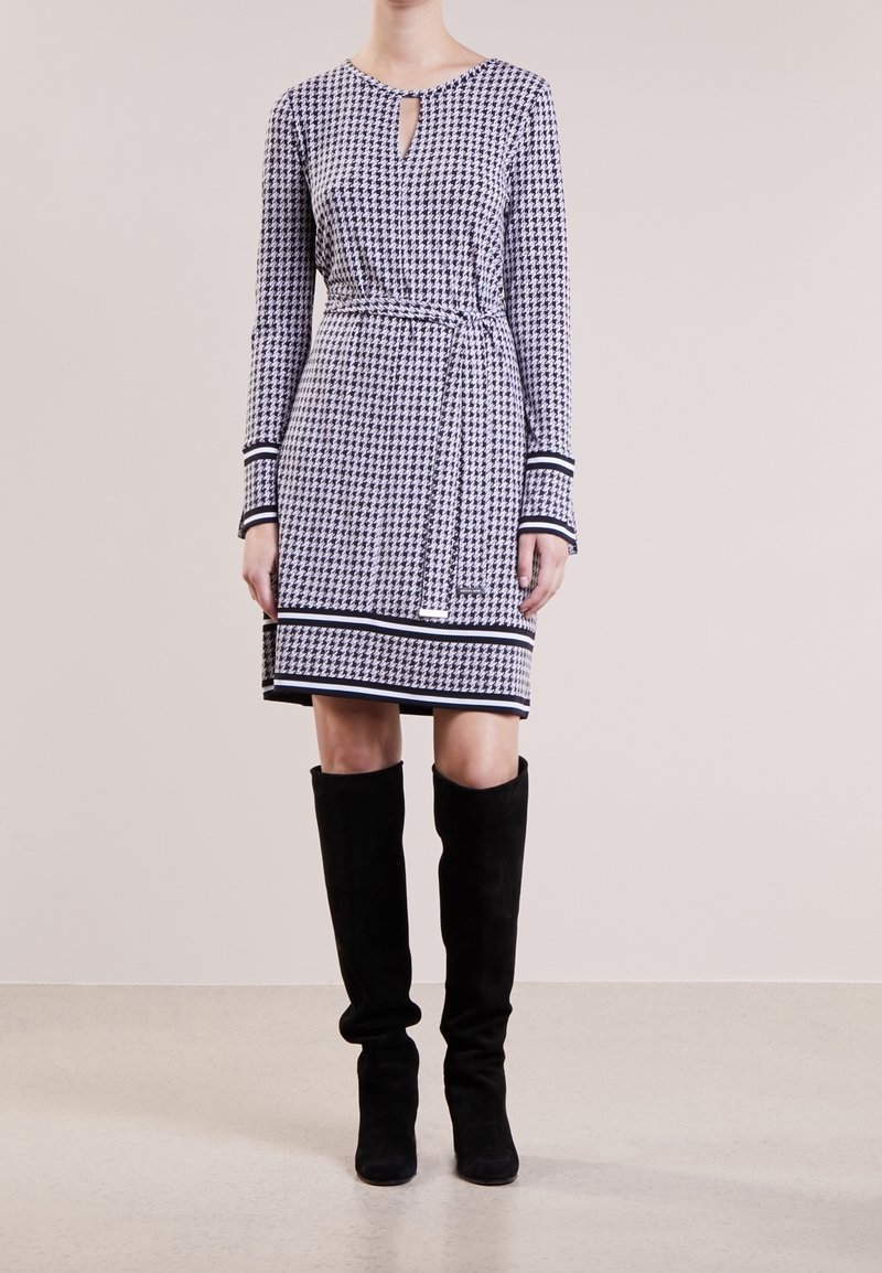 Robe pied-de-poule noire et blanche avec un décolleté en trou de serrure, des manches longues et une taille ceinturée, complétée par des bottes noires montantes jusqu'aux genoux.