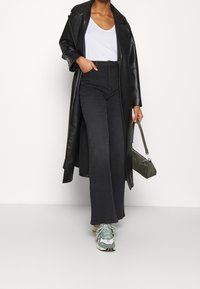 Person wearing black leather long coat, white shirt, high-waisted wide-leg black jeans, green and white sneakers, holding green handbag.