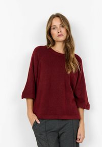 Pulover tricotat burgund cu guler rotund și mâneci trei sferturi, având o croială lejeră și o textură moale. Asortat cu pantaloni gri.