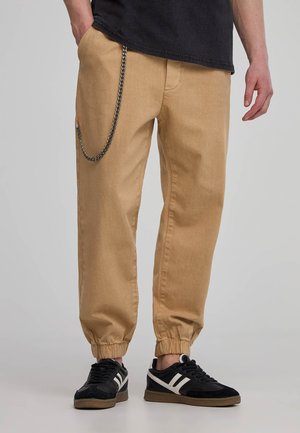 Pantaloni cargo beige con polsini elasticizzati, dotati di una tasca laterale e un accento in catena argentata. Indossati con sneaker nere.