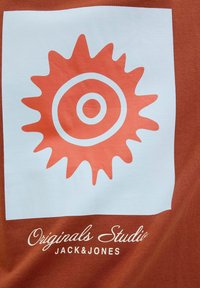 Brun t-shirt med en hvid firkant med en orange solstråle-design og koncentriske cirkler. Teksten nedenfor skriver "Originals Studio JACK&JONES."