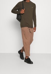 Olivgrüner Strickpullover mit gerippten Bündchen; kombiniert mit braunen Cordhosen; trägt schwarze Schnürschuhe; trägt eine schwarze Tasche.