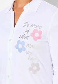 Weißes Hemd mit Knopfverschluss, das bedruckten Text und drei Blumen in Pink, Blau und texturiertem Beige zeigt. Weicher Stoff mit Kragen.