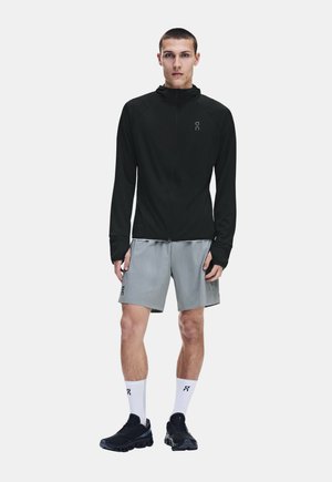Jeune homme debout portant une veste noire zippée, un short gris, des chaussettes blanches montantes et des chaussures de sport noires sur un fond uni.