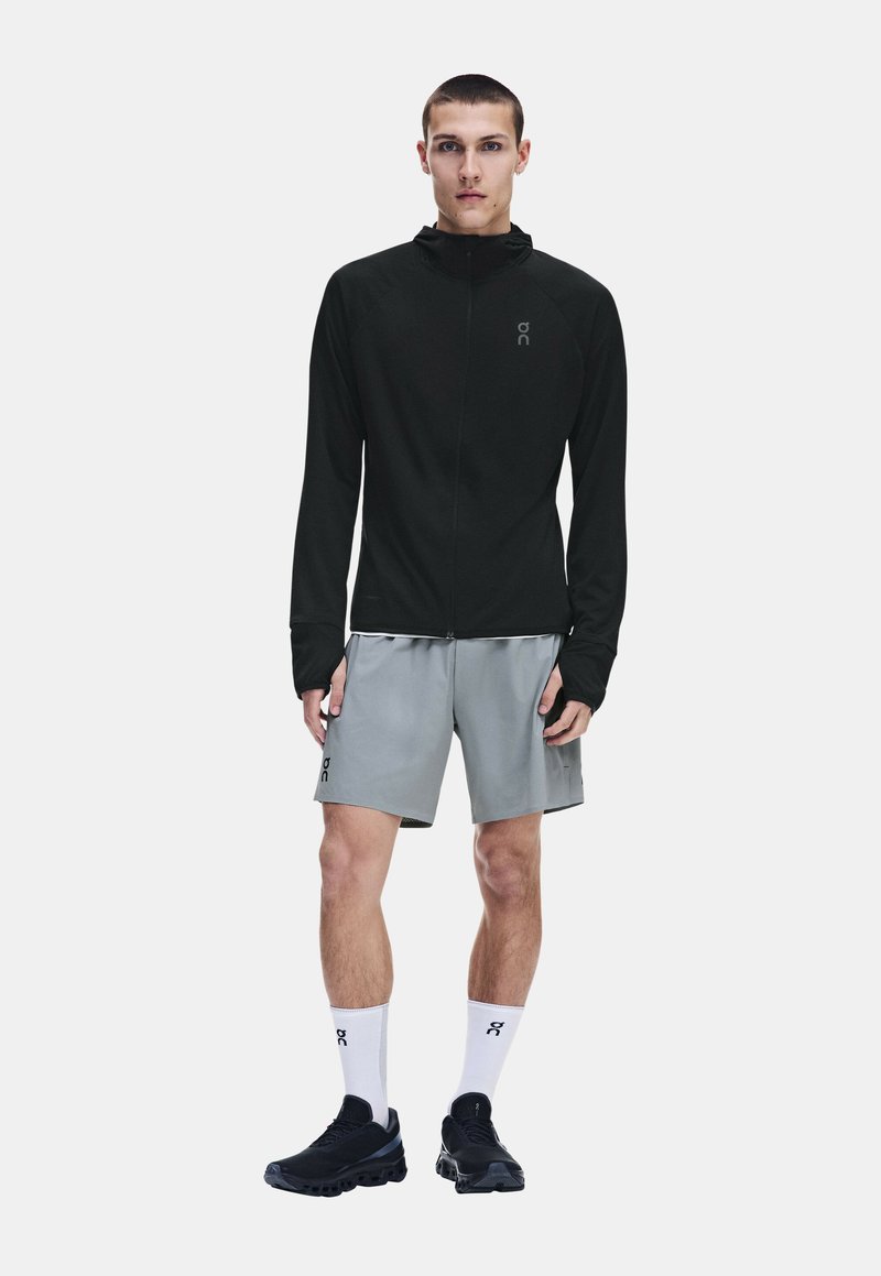 Jeune homme debout portant une veste noire zippée, un short gris, des chaussettes blanches montantes et des chaussures de sport noires sur un fond uni.
