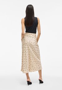 Bež krilo s pikčasto vzorcem iz mehkega, padajočega materiala in srednje dolžine, v kombinaciji s črnim brezrokavnikom in črnimi čevlji s peto.