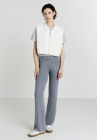Canottiera bianca corta con zip frontale, t-shirt grigia sotto, pantaloni grigi a palazzo e sneakers bianche con accenti marroni. Sfondo neutro.
