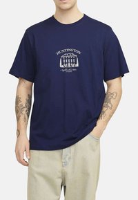 T-shirt en coton bleu marine à manches courtes, avec un graphique blanc d'un bâtiment et le texte "HUNTINGTON" sur le devant.