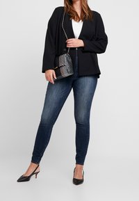 Blazer nero sopra un top bianco, abbinato a jeans skinny scuri e tacchi a punta neri. Piccola borsetta a quadri con tracolla a catena.