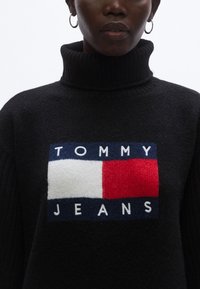 Musta neulottu pooloneule, jossa etuosassa suuri, värikäs logo, jossa sinisiä, valkoisia ja punaisia lohkoja ja teksti "TOMMY JEANS".