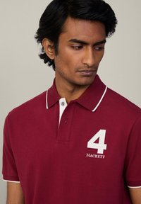 Bordový polo tričko s bielym golierom má vyšívané číslo "4" a logo "HACKETT" na hrudi, vyrobené z bavlnenej tkaniny.