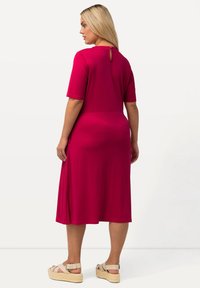 Fuksia, mekko, joka ulottuu polveen, lyhyillä hihoilla ja avointa selkää korostavalla yksityiskohdalla. Valmistettu sileästä kankaasta, jossa on rento istuvuus. Yhdistetty kevyisiin korkokenkiin.