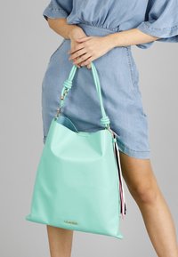 Tote bag v mintové zelené barvě s matným povrchem vyrobený ze syntetického materiálu. Disponuje uzlovým popruhem a barevnými třásněmi, má obdélníkový tvar a měkkou strukturu.