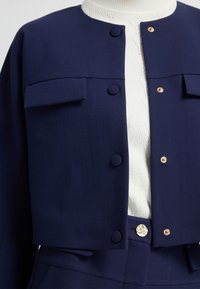 Granatowa krótka kurtka z dwoma kieszeniami na piersi, okrągłym dekoltem i złotymi akcentami. Noszona na teksturowanym białym topie. Gładka tkanina.