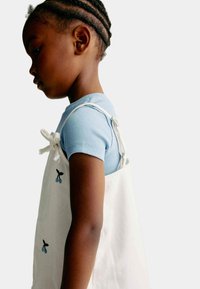 Hvid forklæde-lignende kjole med blå kirsebærembroderi, båret over en lyseblå ribbet kortærmet top. Har justerbare bindebånd ved skuldrene.