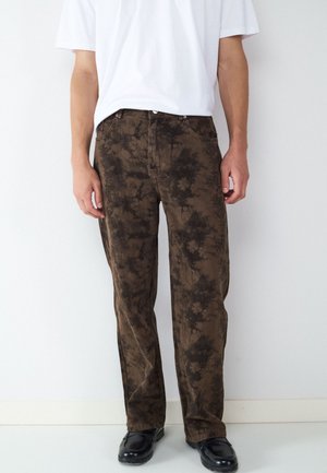 Hombre que lleva pantalones con patrón tie-dye marrón y negro, camisa blanca de manga corta y zapatos negros, de pie contra una pared blanca.
