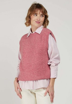 Femme aux cheveux courts et ondulés portant un gilet sans manches en tricot rose par-dessus une chemise boutonnée rayée rose et blanche et un pantalon de couleur claire.
