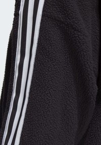 Svart fleecejacka med en texturerad yta, med tre vita ränder längs ärmen gjorda av vävt tyg.