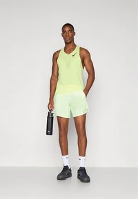 Světlé zelené sportovní tílko bez rukávů s designem z síťoviny, k němu světle zelené šortky, černá láhev na vodu a černé tenisky.