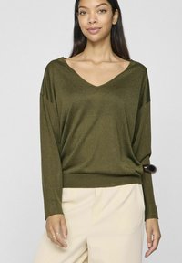 Oliwkowy, oversize sweter z dekoltem w serek, z długimi rękawami, prążkowanymi mankietami i dołem. Gładka struktura, luźno opada na parę jasnobeżowych spodni.