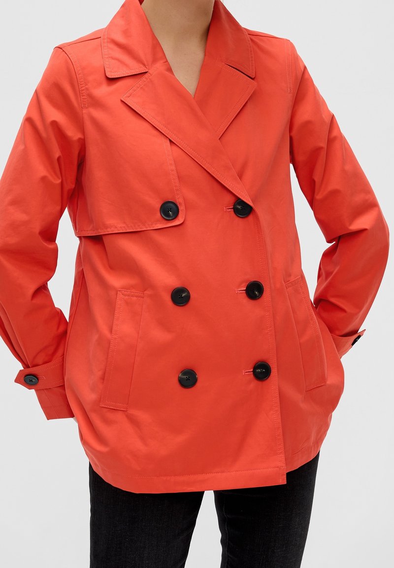 Personne portant une veste orange vif à double boutonnage avec des boutons noirs et des poches latérales, les mains dans les poches, sur un fond uni.