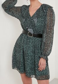 Vestido de leopardo verde con mangas largas y transparentes, escote con volantes y cinturón negro en la cintura. Tejido ligero y fluido.