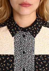 Krage skjorta med en kombination av blommönster i svart, rosa och gult. Har knappar och olika texturerade tygpaneler.