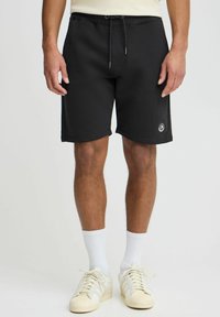 Shorts de sport noirs avec une taille élastique et un cordon de serrage, comportant un petit logo en bas à gauche. Texture lisse.