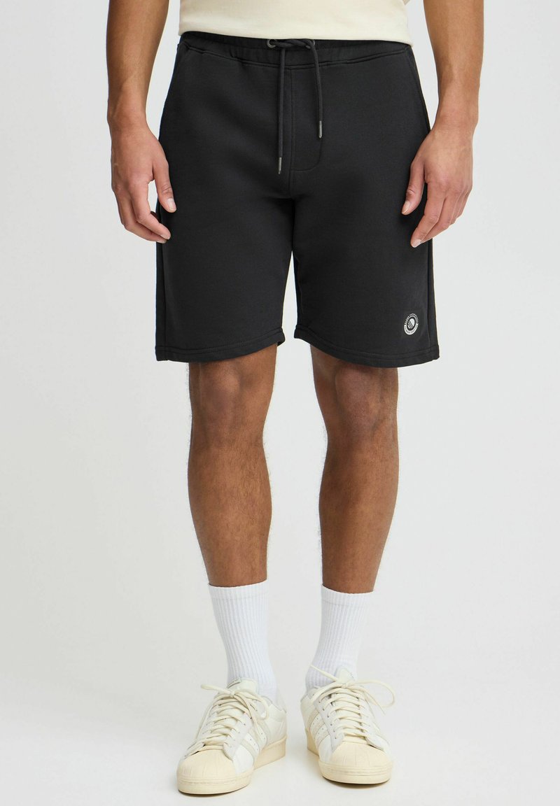 Shorts de sport noirs avec une taille élastique et un cordon de serrage, comportant un petit logo en bas à gauche. Texture lisse.