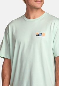 Světle mátově zelené bavlněné tričko s krátkými rukávy. Obsahuje malé vícolorové grafické logo na levém hrudníku, hladkou texturu a volný střih.