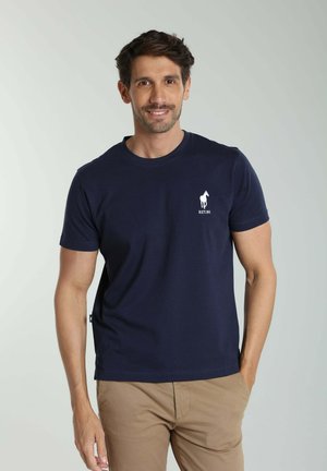 Homme souriant aux cheveux foncés portant un t-shirt bleu marine avec un logo de cheval blanc et un pantalon beige, debout avec une main dans la poche.