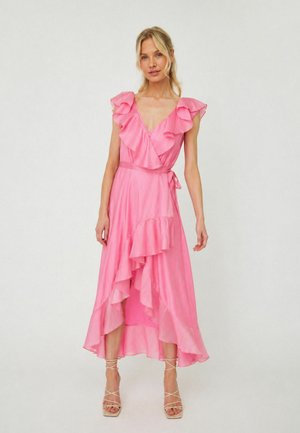Donna che indossa un vestito rosa senza maniche a portafoglio con volant e tacchi beige con cinturini, in piedi davanti a uno sfondo chiaro uniforme.