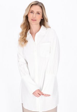 Femme blonde aux ondulations lâches portant une chemise blanche oversize à manches longues avec une poche avant, se tenant devant un fond blanc.