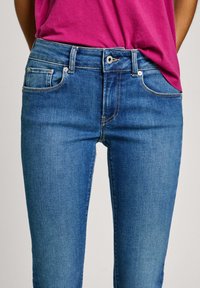 Sinine denimist teksapüksid on sobiva lõikega, keskmise tõusuga vöökohaga, esi-taskute ja kergelt õmmeldud detailidega. Koos roosa t-särgiga.