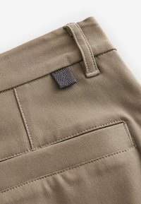 Brauner Baumwollstoff mit glatter Textur, versehen mit einer Gesäßtasche und einem kleinen Stoffetikett. Die Nähte sind entlang der Ränder sichtbar.