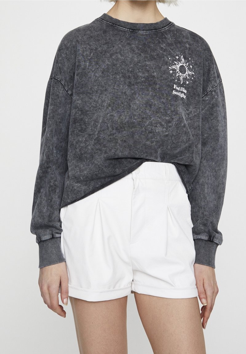 Personne portant un t-shirt à manches longues gris foncé avec un motif de soleil et le texte "Feel like Sunlight", assorti à un short blanc taille haute.