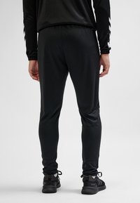 Mężczyzna w czarnych sportowych spodniach, czarnych trampkach i czarnej bluzie z długim rękawem, stojący tyłem na jednolitym szarym tle.