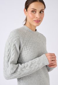 Maglione lavorato a maglia di un grigio chiaro con collo rotondo; presenta motivi a trecce sulle maniche e una texture a coste sul corpo. Tessuto morbido.