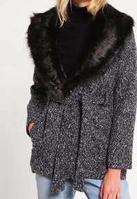 Abrigo de manchas en blanco y negro con un gran collar de piel sintética, que presenta una cintura con cinturón, dos bolsillos frontales y un diseño de tejido texturizado.