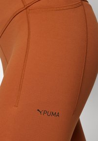 Narančaste tajice izrađene od glatkog, rastezljivog materijala s prilagođenim oblikom. Sadrže crni Puma logo i vidljive detalje šavova.
