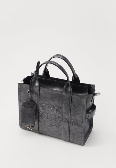 Zwarte leren tote bag met reliëftekst, stevige handvatten en een afneembare label. Beschikt over een gestructureerde vorm en een textuurafwerking.